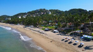 A beach with rows of sun loungers and umbrellas, a clear sky, and buildings in the background. - Casa Palmar 3BR  · Remarkable 3BR, 2BA Beachfront Condo (Sayulita)