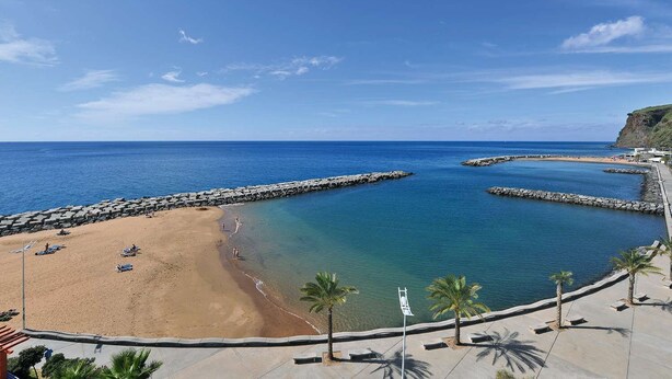 A beach with a rocky breakwater, clear blue water, and palm trees. - Modern Madeira house in Calheta, Madeira (Estreito da Calheta)
