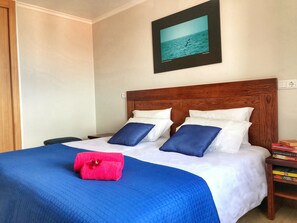 Ein ordentlich bezogenes Bett mit einer blauen Steude, weißen Kissen und einem roten Handtuch. Über dem Bett hängt ein gerahmtes Bild vom Meer.