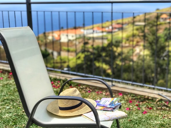 A chair with a straw hat and travel brochures on a grassy area with a view of a coastal landscape. - Modern Madeira house in Calheta, Madeira (Estreito da Calheta)