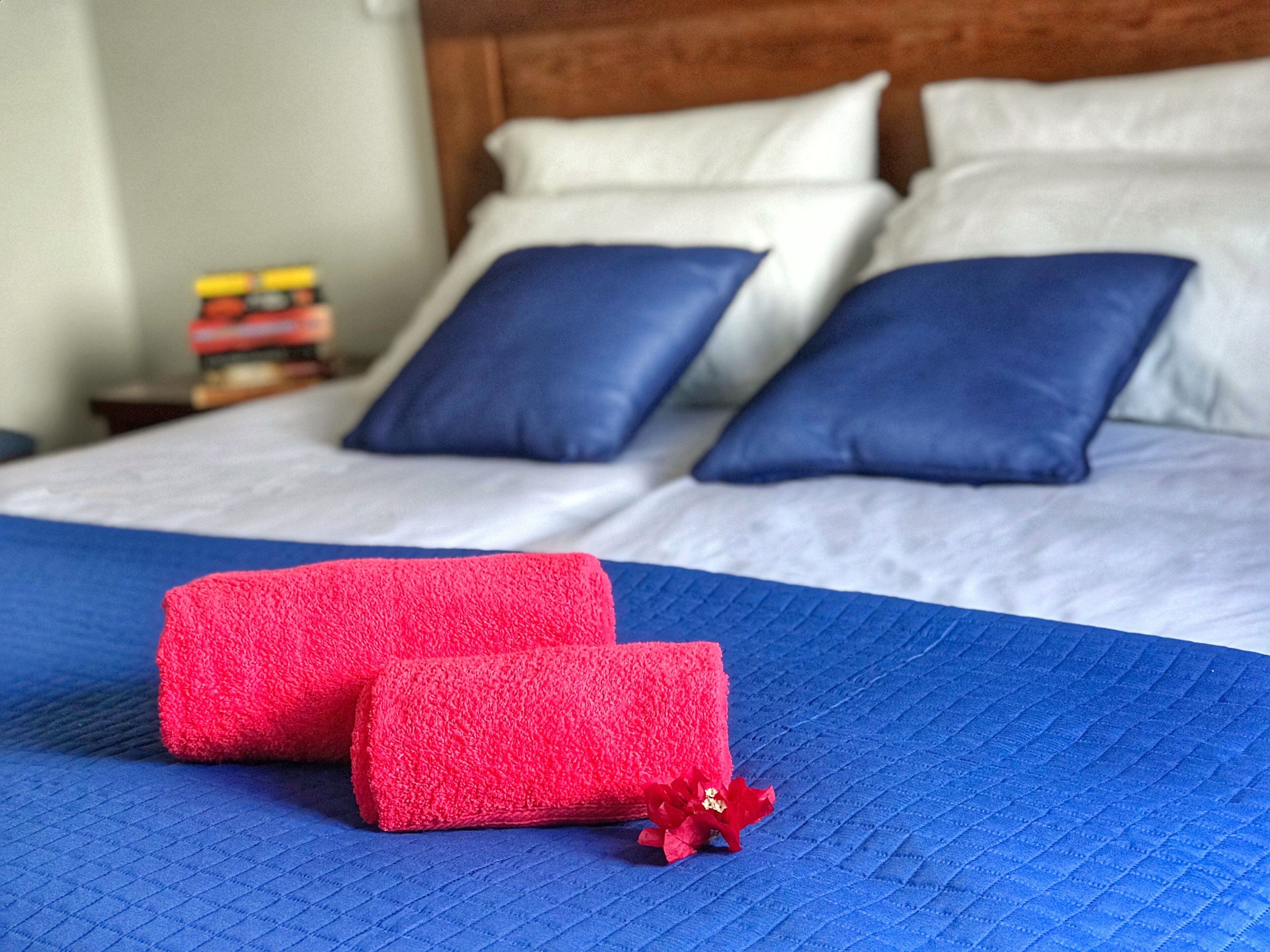 Una cama bien tendida con ropa de cama azul y blanca, dos toallas rojas y una pequeña flor decorativa.