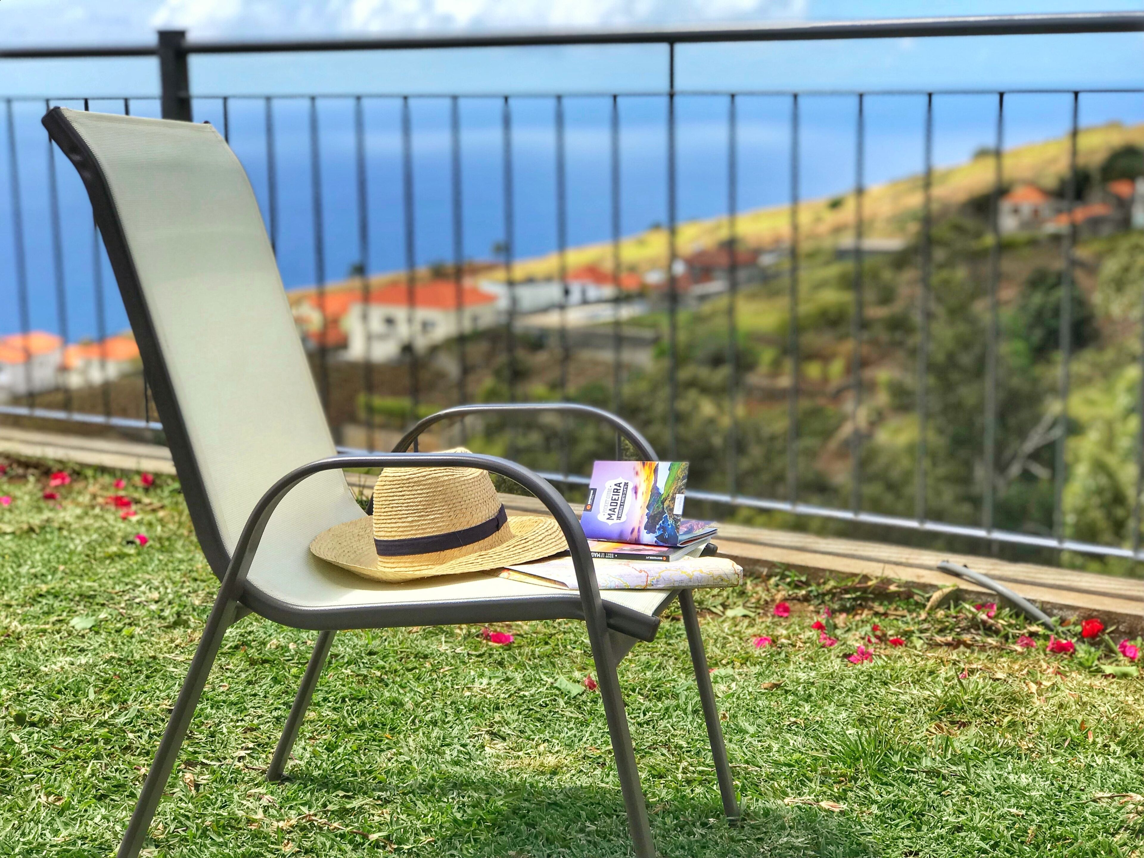 A chair with a straw hat and a book on a grassy area with a railing and a view of a town and hills in the background.