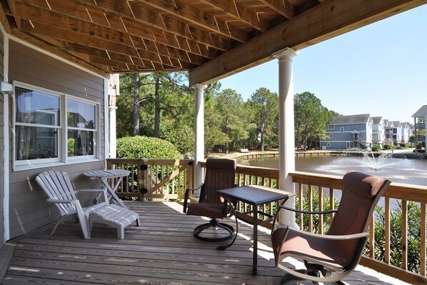 A wooden deck with outdoor furniture, a view of a water body, and residential buildings in the background. - The Bates Motel: Pet friendly, Oceanside Condo (Corolla)