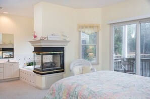 Ein Schlafzimmer mit Kamin, einem großen Fenster mit Aussicht, einem weißen Sessel und einem Bett mit blumigem Überwurf.