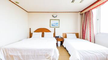 A hotel room with two single beds, a wooden headboard, a nightstand, a small table, a framed picture on the wall, and a window with curtains.