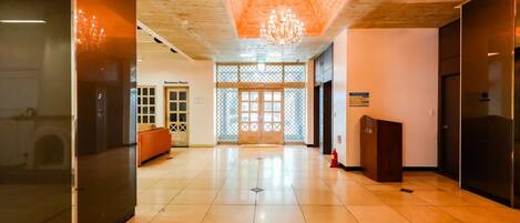 Um hall espaçoso com teto de madeira, um candelabro, um balcão de recepção e uma sala de negócios visível ao fundo.