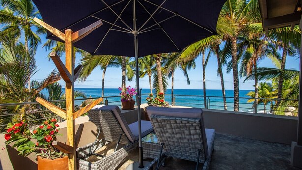 A balcony with wicker chairs, a large umbrella, and a view of the ocean and palm trees.