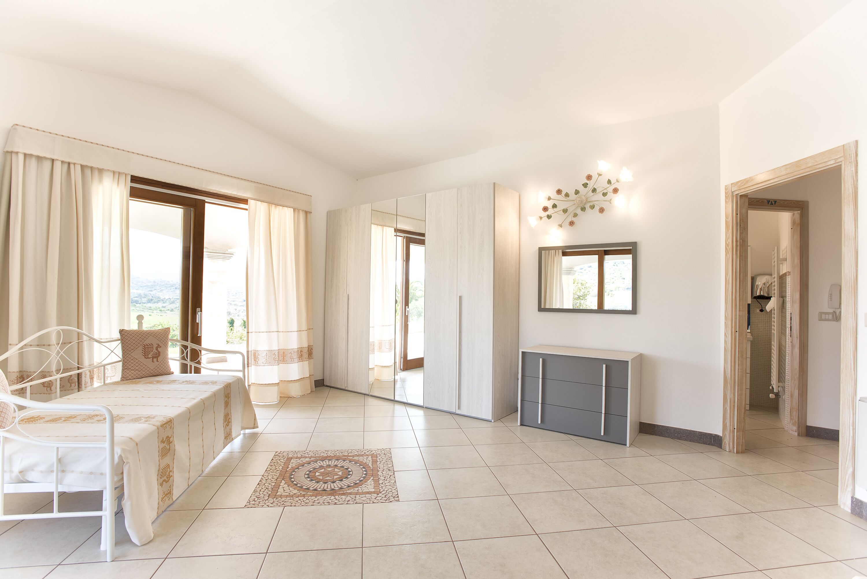 A spacious bedroom with a bed, a bedside table, a mirror, and a view of the outdoors.