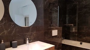 Un baño moderno con detalles de mármol en la pared, un espejo redondo, un lavabo blanco, un inodoro y una zona de ducha con mampara de vidrio.