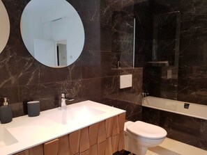 A modern bathroom with a marble backsplash, a round mirror, a white sink, a toilet, and a glass-enclosed shower area.