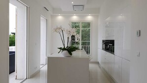 Una cocina moderna con armarios blancos, una isla central con una orquídea en maceta y vistas a través de la puerta de vidrio.