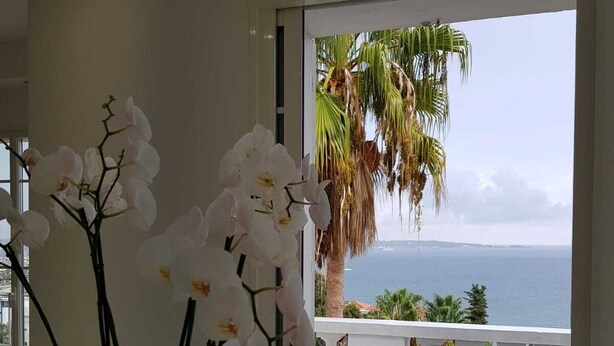 Habitación moderna con un ventanal que ofrece vistas al mar, con orquídeas blancas sobre una mesa y un balcón con mobiliario exterior.