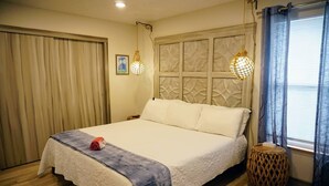A neatly made bed with a white comforter and a blue blanket, a wooden floor, a woven stool, and a window with blue curtains. - Fall Specials Available! Pet Friendly! Steps to white sandy beaches and the bay!  Discounts applied for stays longer than 7 days and 30 days! (Bradenton Beach)