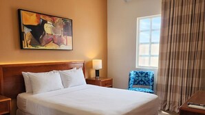 Ein Hotelzimmer mit einem großen Bett, einem Nachttisch, einer Lampe, einem Sessel, einem Schreibtisch, einem Fenster mit Vorhängen und einem Bild an der Wand.