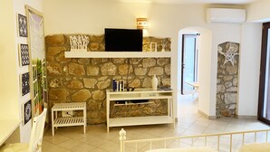 A room with a stone wall, a flat-screen TV, a small table with books, a white chair, and a bed with white bedding. - Circeii, apartment with bedroom, living room and exterior with bathtub (San Felice Circeo)