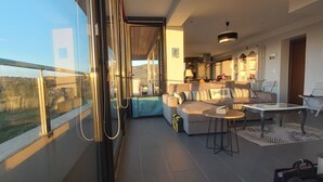 Een balkon met glazen hekwerk, een bank, een kleine tafel en uitzicht op een bergachtig landschap.