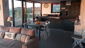 Een moderne woonkamer met een bank, eettafel en stoelen. Achtergrond toont een keuken met een koelkast, oven en spoelbak.
