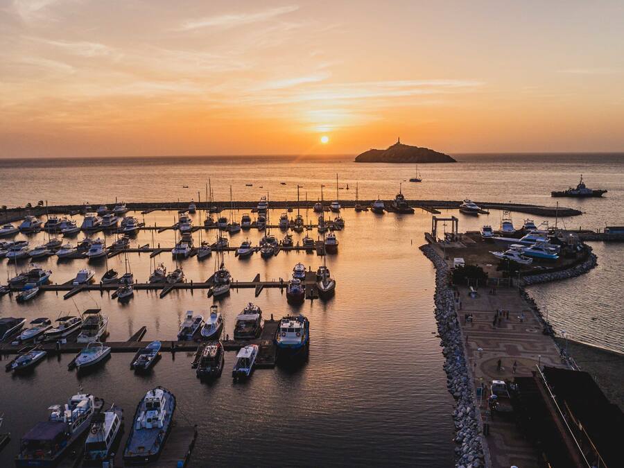 Una marina con barcos amarrados en muelles, un muelle con gente y un atardecer al fondo.