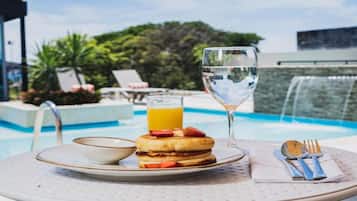Une table dressée pour le petit-déjeuner avec une assiette de crêpes, un verre de jus d’orange, un verre d’eau et un bol, le tout placé sur une table près d’une piscine.