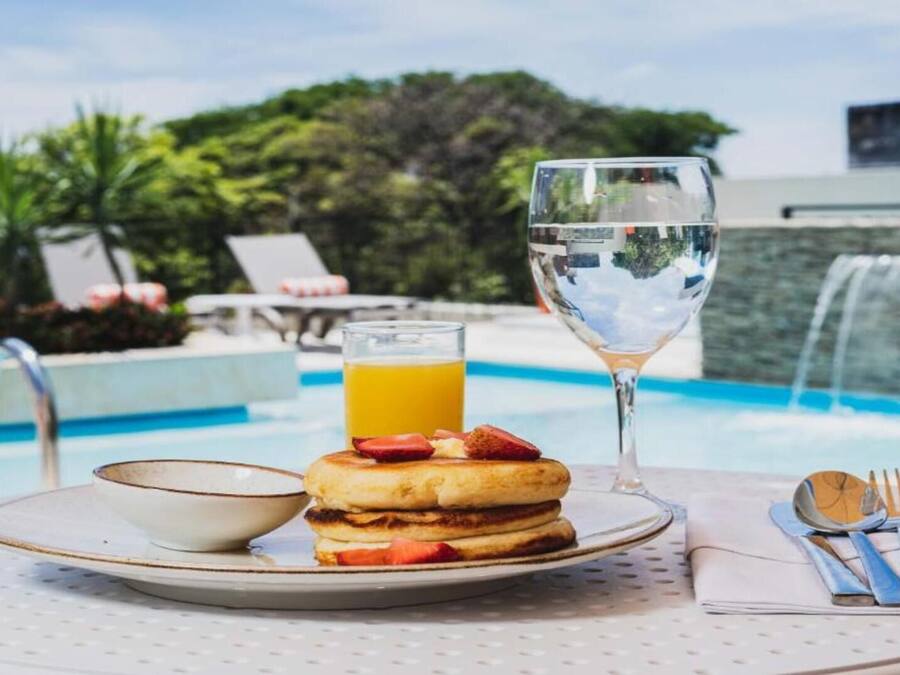 Une table dressée pour le petit-déjeuner avec une assiette de crêpes, un verre de jus d’orange, un verre d’eau et un bol, le tout placé sur une table près d’une piscine.