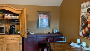 Una cucina funzionale con mobili in legno, un forno a microonde e una macchina da caffè. Una cornice con un poster decora la parete, e un tavolo con sedie è posizionato al centro.