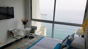 Une chambre d’hôtel avec un grand lit, une petite table avec des chaises, un téléviseur fixé au mur et une vue sur la mer par de grandes fenêtres.