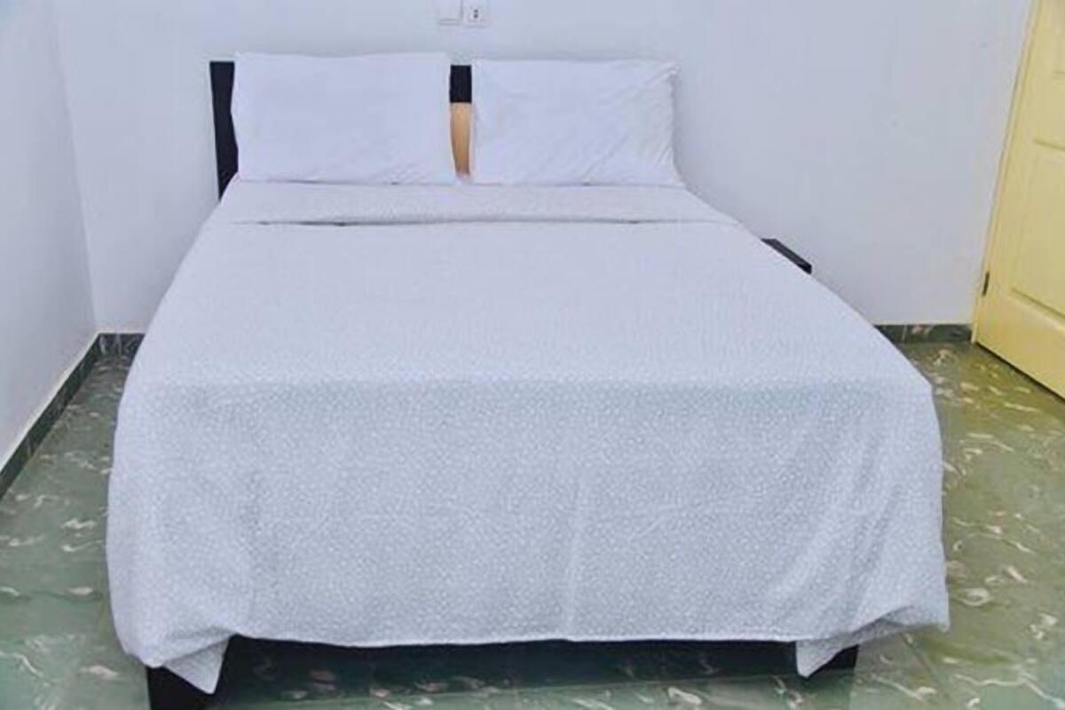 A single bed with a white bedspread and two pillows in a room with white walls and a green marble floor.