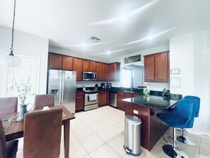 A modern kitchen with wooden cabinets, stainless steel appliances, a dining area with a table and chairs, and a trash can.