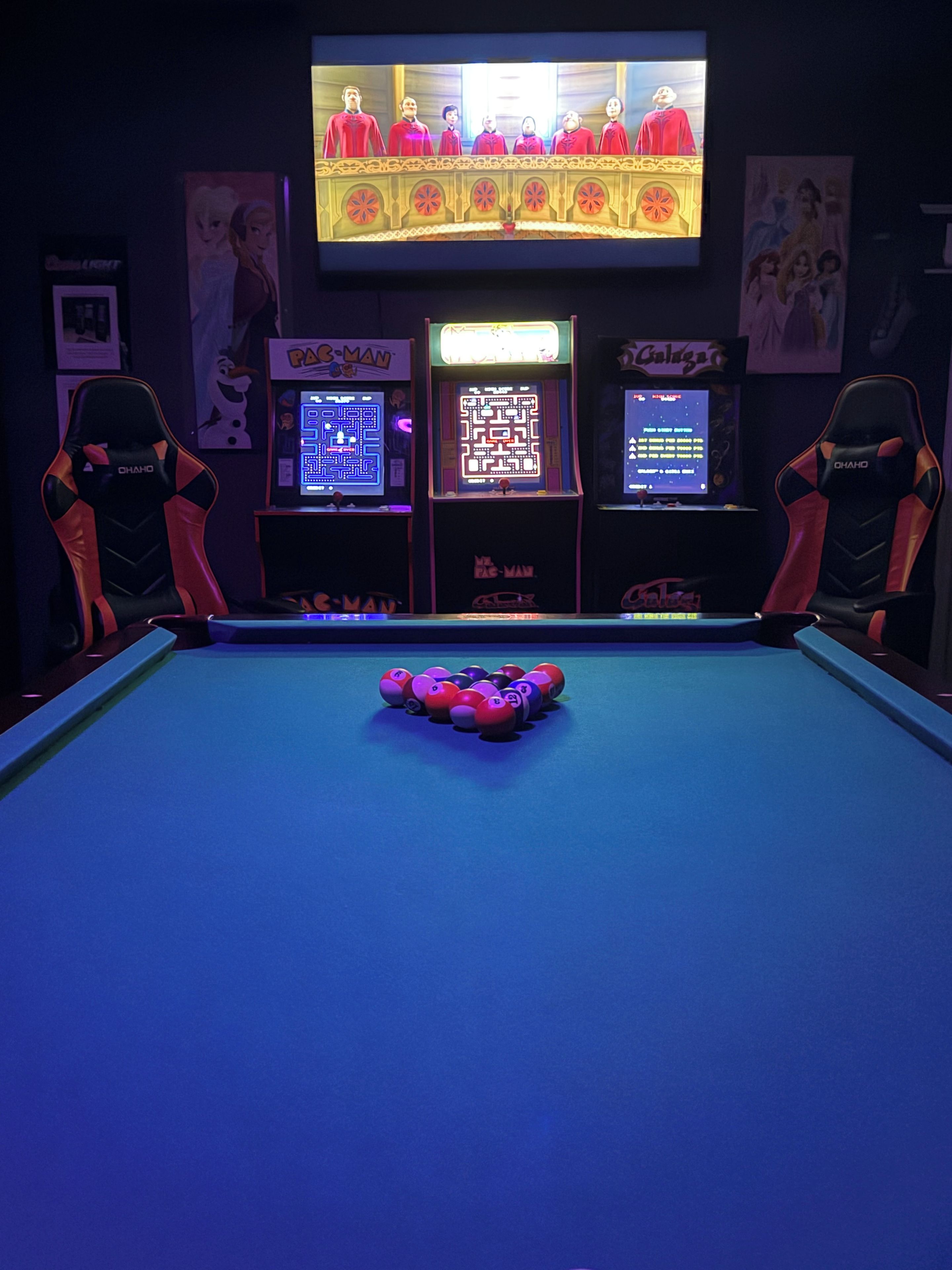 Een pooltafel met een keu en ballen, aan weerszijden arcadekasten en twee game stoelen.