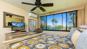 A hotel room with a bed, a TV, a ceiling fan, a window with a view of palm trees and the ocean, and a dresser.