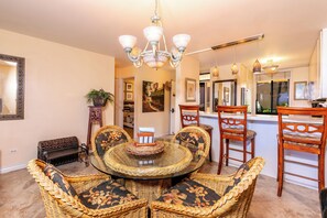 A dining area with a round glass table surrounded by wicker chairs, a chandelier, and a kitchen area with wooden chairs and a bar.