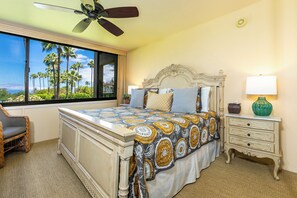 A bedroom with a large window, a bed with patterned bedding, a nightstand, a chair, and a ceiling fan.