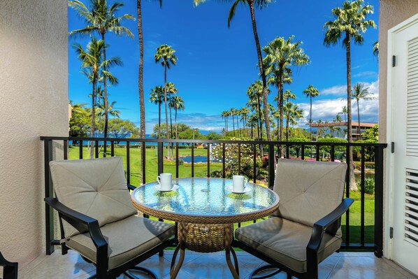 A balcony with two chairs and a table, overlooking a scenic view of palm trees and a golf course.