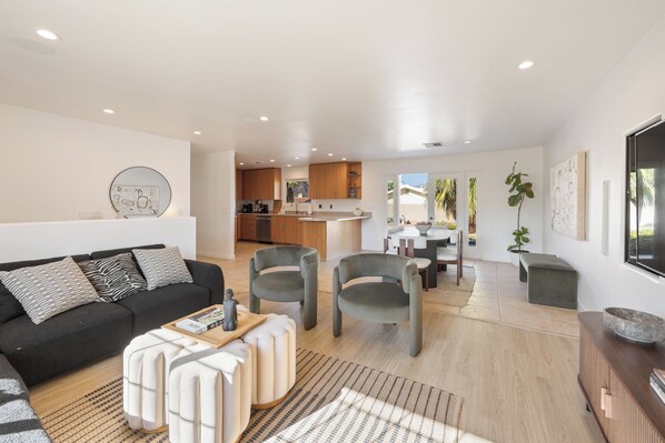 A modern living room with a black sofa, grey armchairs, and a coffee table. A kitchen with wooden cabinets and a dining area with a table and chairs are visible in the background.