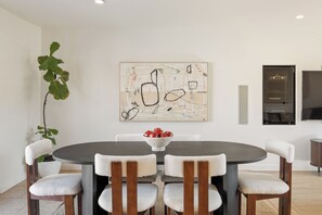 A dining area with a round table, six chairs, a bowl of fruit, and a large abstract painting on the wall.