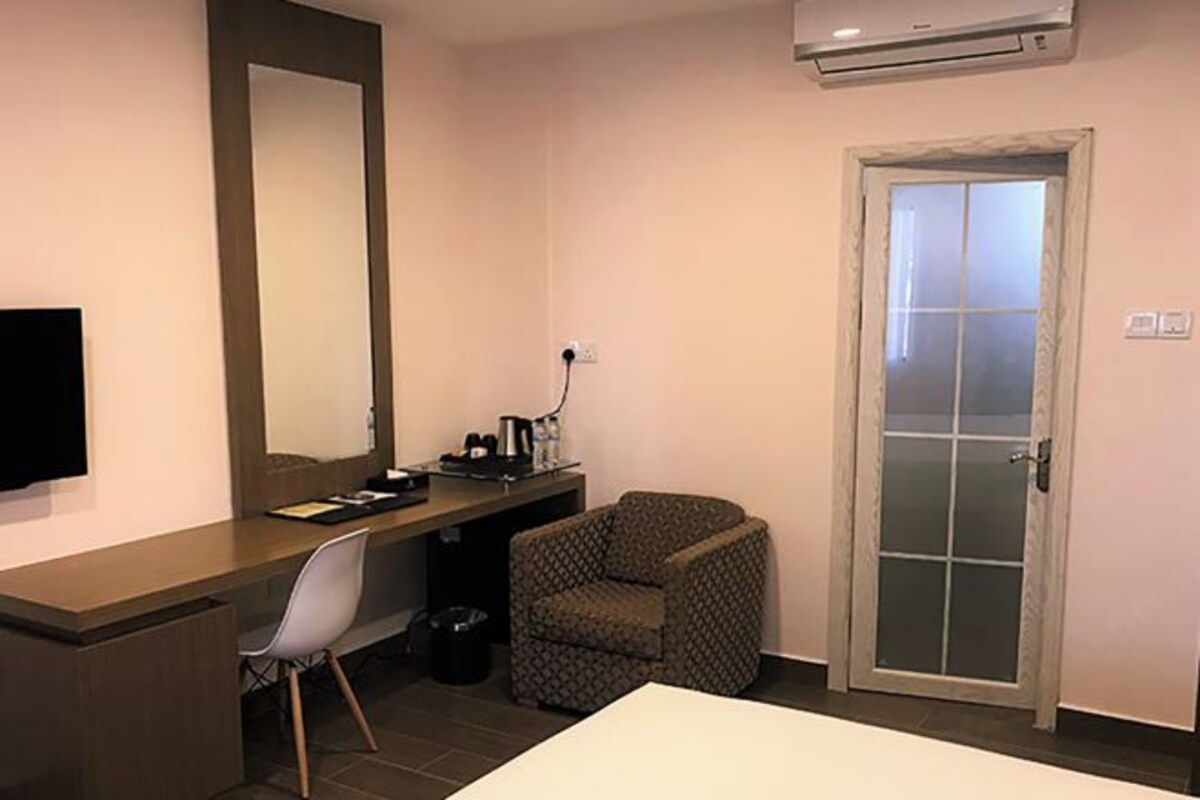 A hotel room with a bed, a desk with a chair, a small armchair, a mirror, a door with a frosted glass panel, and an air conditioning unit.