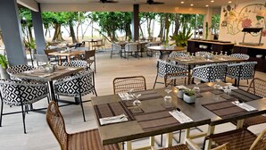 Un vaste espace extérieur aménagé pour les repas, avec de nombreuses tables et chaises, entouré de grandes fenêtres qui offrent une vue sur les arbres et une plage.