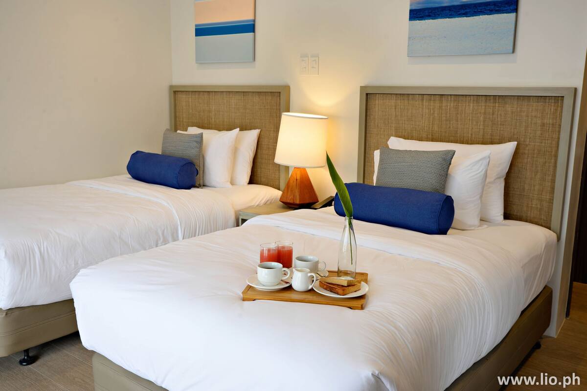 Ein Hotelzimmer mit zwei Betten, einem hölzernen Nachttisch, einer Lampe, einem Tablett mit Frühstücksgegenständen und Kunstwerken an den Wänden.