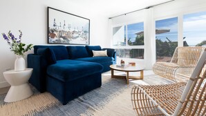 A modern living room with a blue sofa, a wicker chair, a white side table, and a large window offering a view of the outdoors. - Avalon 2 Unit Buyout (I & III) AvantStay | Mins to Gaslamp| Beautiful Deck w/BBQ (San Diego)