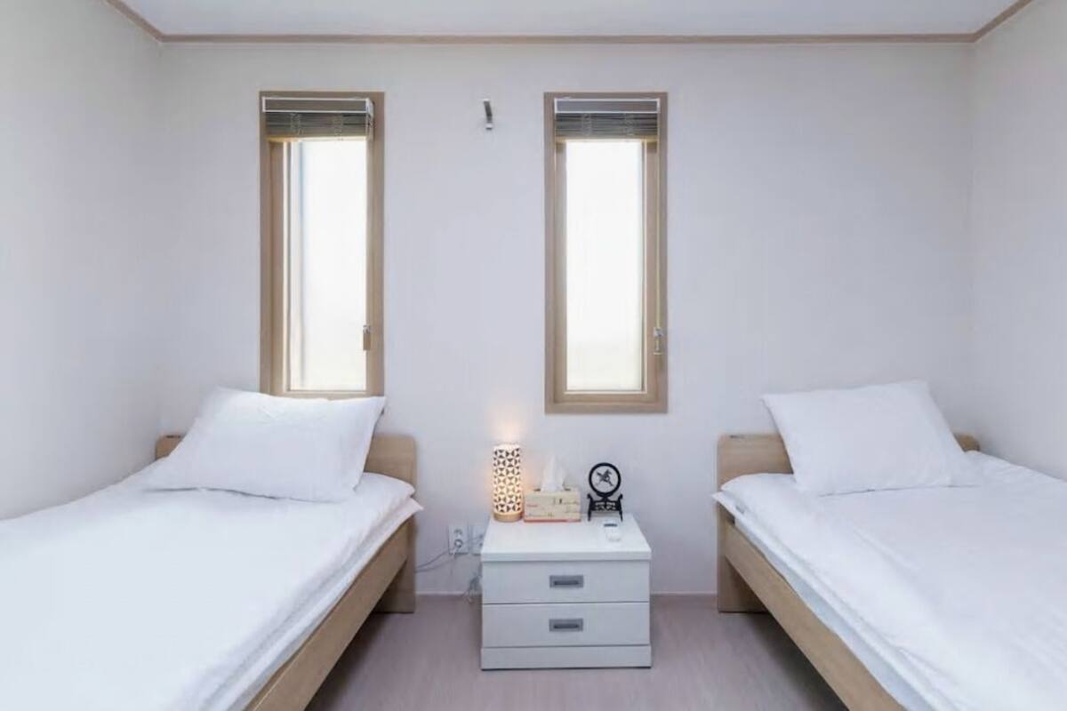 A bedroom with two single beds, a bedside table with a lamp, and two windows.