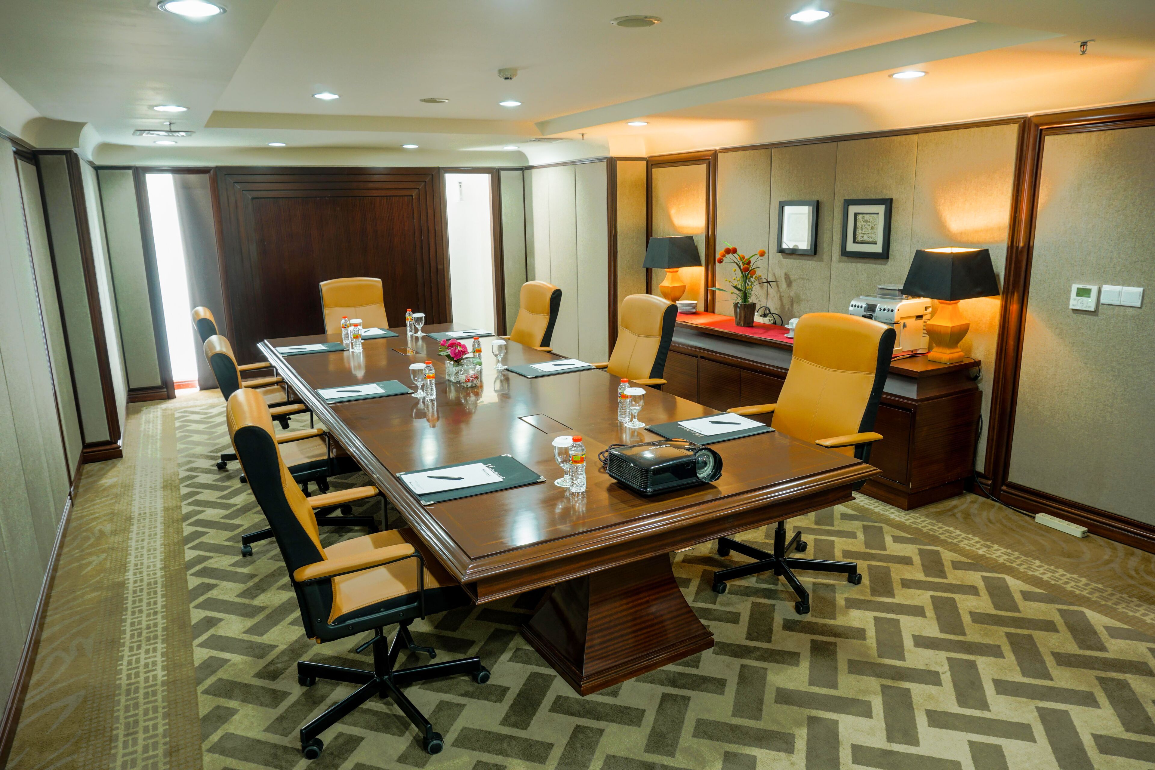 Un salón de conferencias con una larga mesa, varias sillas, un proyector y una mesa pequeña con una lámpara y flores.