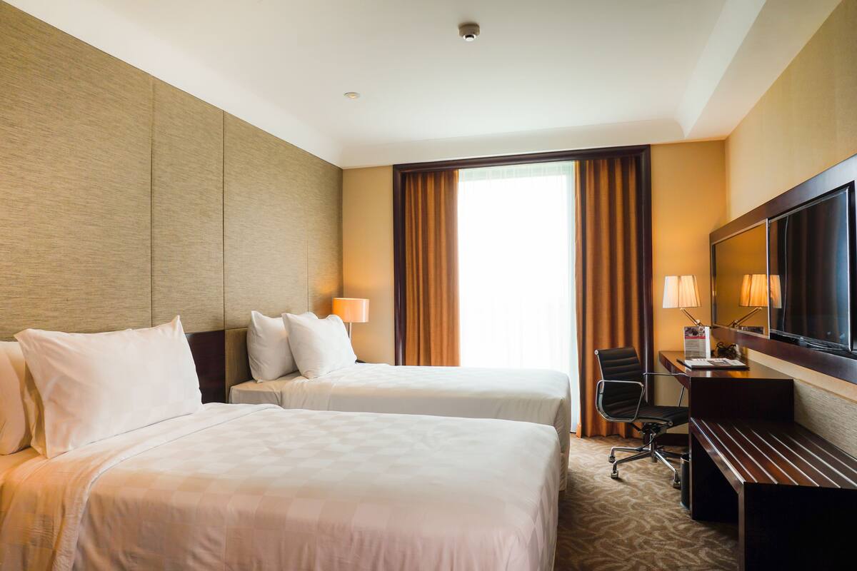 Une chambre d’hôtel avec deux lits, un bureau, une chaise, une télévision et une grande fenêtre avec des rideaux.