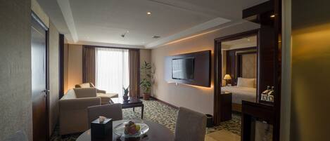 Habitación de hotel con una mesa de comedor, dos sillones, un sofá, un televisor montado en la pared y una cama con una mesita de noche.