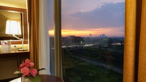 Une chambre d’hôtel avec vue sur les toits d’une ville au crépuscule, une table en bois ornée d’un vase rempli de fleurs roses et un miroir qui reflète la lampe et la fenêtre.