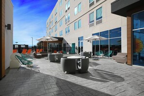 Un área de estar moderna al aire libre con mesas y sillas, cielo despejado y un edificio con amplios ventanales al fondo.