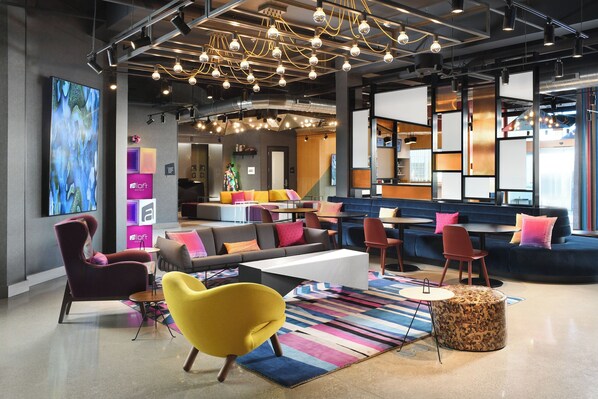 A modern lounge area with seating arrangements, a large flat-screen TV, and a colorful striped rug. - Aloft by Marriott Dallas Euless (Euless)