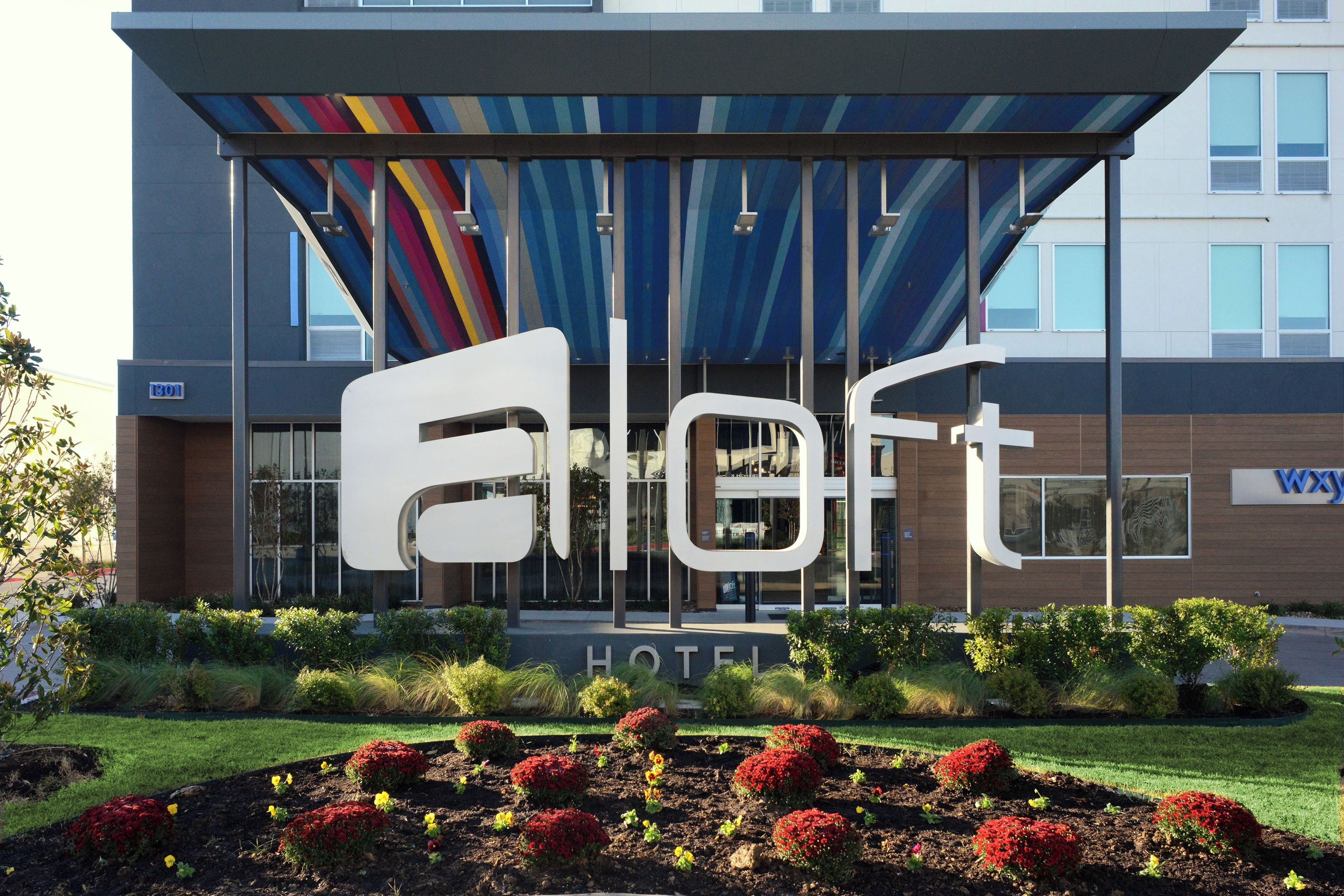 A hotel with a modern facade, a large sign with the name 'Eloft', and a well-maintained garden with red flowers.