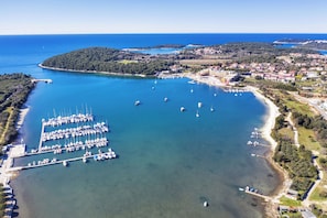 An aerial view of a coastal marina with numerous boats docked, surrounded by a small town with buildings and greenery.