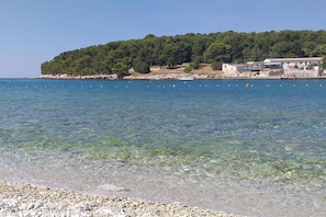 Eine Küstenlandschaft mit klarem Wasser, einem felsigen Strand und einem bewaldeten Hügel mit Gebäuden im Hintergrund.