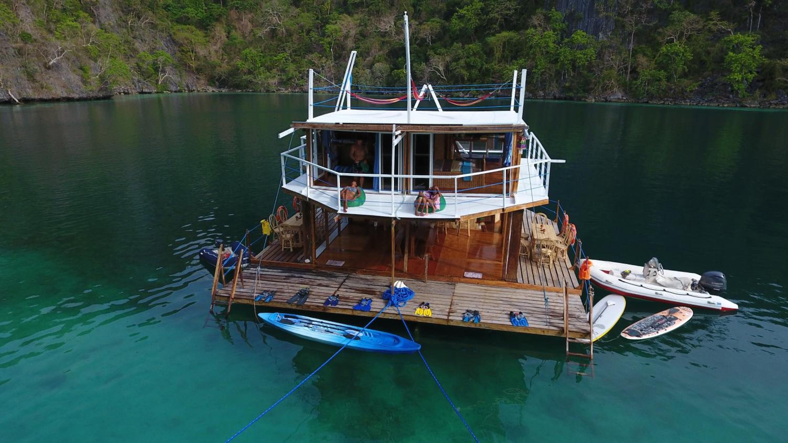 A boat with a wooden deck, a covered upper deck, and a small cabin. There are two kayaks and a surfboard on the deck.
